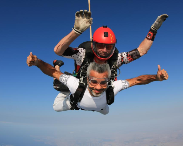 Umar Farooq Zahoor mid-air during a thrilling tandem skydiving experience, smiling  against a backdrop of clear blue skies.